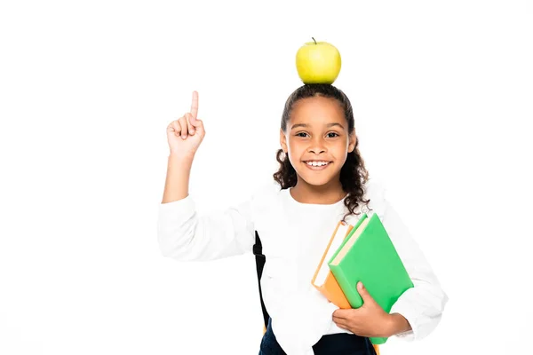 Cheerful african american schoolgirl with apple on head holding books and showing idea gesture isolated on white — Stock Photo
