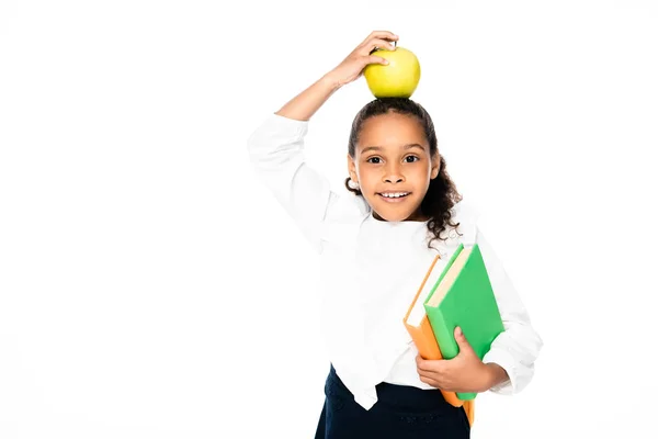 Adorable african american schoolgirl holding apple on head while standing with books isolated on white — Stock Photo