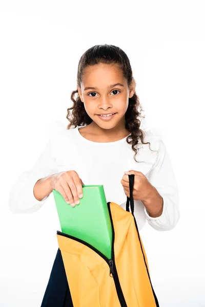 Усміхнена афроамериканська школярка кладе книгу в рюкзак ізольовано на білому — стокове фото
