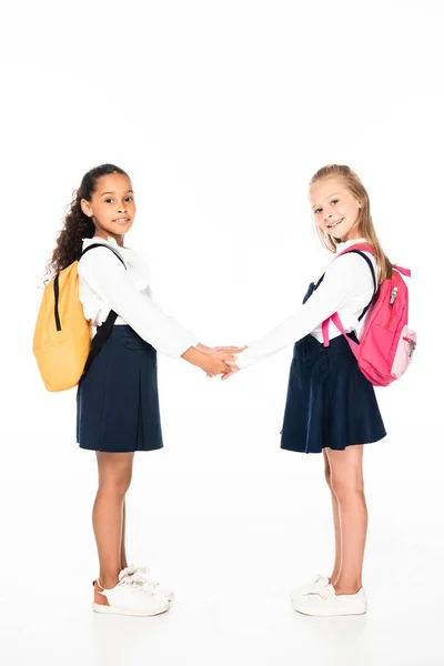 Volle Länge Ansicht von zwei niedlichen multikulturellen Schulmädchen, die Hände halten und in die Kamera auf weißem Hintergrund schauen — Stockfoto