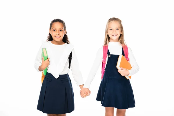 Две очаровательные мультикультурные школьницы держатся за руки и смотрят на камеру, изолированную на белом — стоковое фото