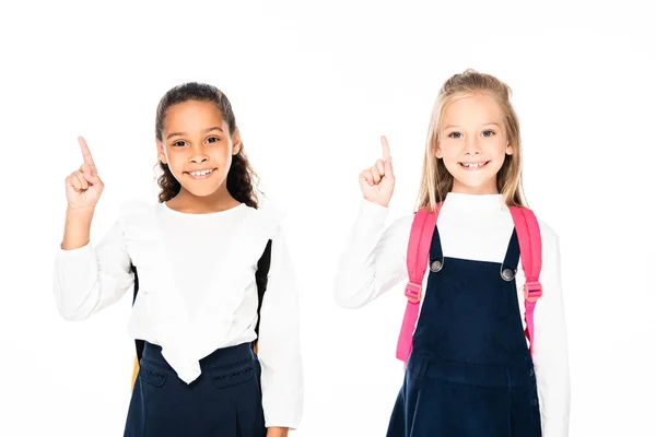 Two cheerful multicultural schoolgirls showing idea gestures while smiling at camera isolated on white — Stock Photo