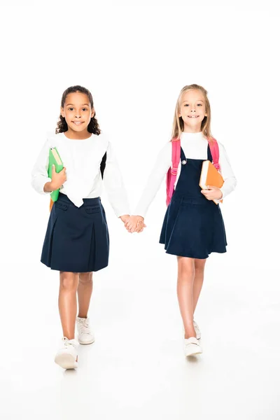 Vista completa de dos colegialas sonrientes y multiculturales caminando mientras se toman de la mano sobre fondo blanco - foto de stock