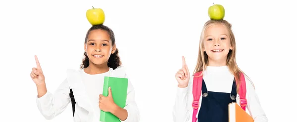 Plano panorámico de dos colegialas multiculturales alegres con manzanas en la cabeza mostrando gestos de idea aislados en blanco - foto de stock