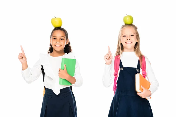 Zwei fröhliche multikulturelle Schulmädchen mit Äpfeln auf dem Kopf, die Ideengesten isoliert auf weiß zeigen — Stockfoto
