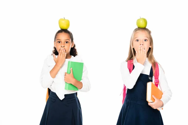 Deux écolières multiculturelles choquées avec des pommes sur la tête couvrant la bouche avec les mains isolées sur le blanc — Photo de stock