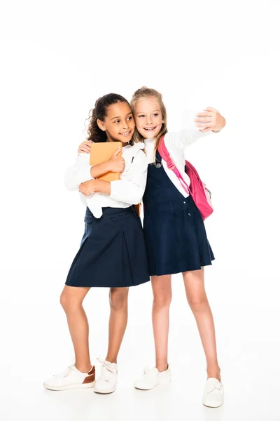 Vista completa de la colegiala feliz abrazando amigo afroamericano y tomando selfie sobre fondo blanco - foto de stock