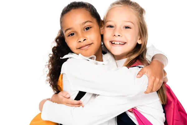 Duas colegiais multiculturais felizes abraçando enquanto sorrindo para a câmera isolada no branco — Fotografia de Stock