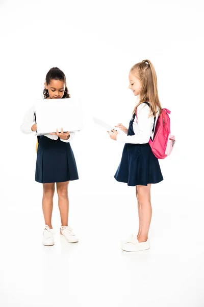 Vue pleine longueur de deux écolières multiculturelles en utilisant ordinateur portable et tablette numérique sur fond blanc — Photo de stock