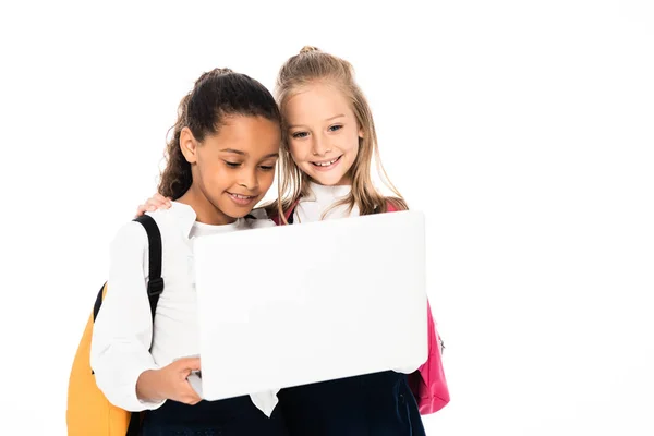 Duas alunas multiculturais alegres sorrindo ao usar laptop isolado no branco — Fotografia de Stock