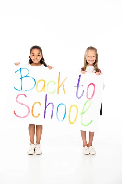 Deux écolières multiculturelles joyeuses tenant une pancarte avec inscription de retour à l'école sur fond blanc — Photo de stock