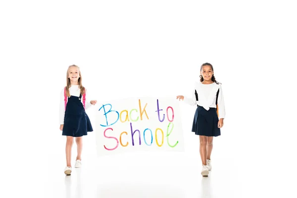 Full length view of two multicultural schoolgirls holding placard with back to school inscription while walking on white background — Stock Photo