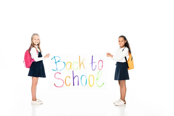 Vista completa de dos colegialas multiculturales con mochilas con pancarta con inscripción de regreso a la escuela sobre fondo blanco - foto de stock