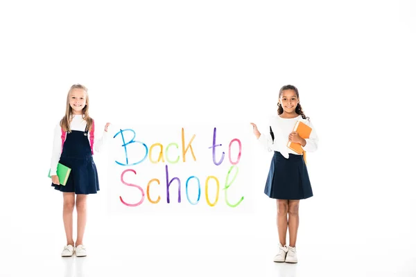 Vista completa de dos colegialas multiculturales sosteniendo libros y pancarta con inscripción de regreso a la escuela sobre fondo blanco - foto de stock