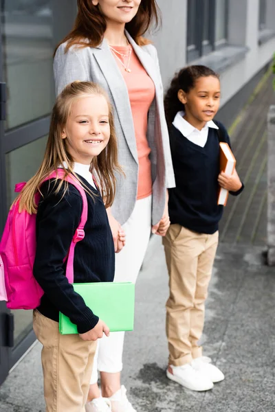 Vista recortada de la madre cogida de la mano de dos hijas multiculturales en uniforme escolar - foto de stock