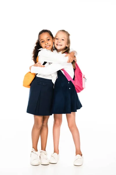 Vista completa de dos colegialas multiculturales felices abrazando y sonriendo a la cámara sobre fondo blanco - foto de stock