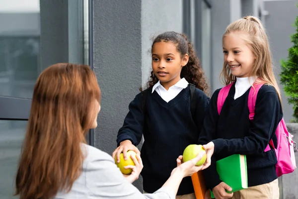 Vista posteriore della madre che dà mele a due figlie multiculturali che vanno a scuola — Foto stock