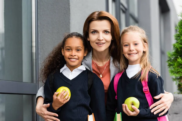 Feliz madre abrazando alegre multicultural hijas y sonriendo a la cámara mientras de pie cerca de la escuela - foto de stock