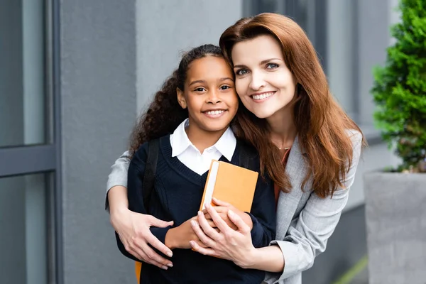 Glückliche Mutter umarmt adoptierte afrikanisch-amerikanische Tochter, während sie in der Nähe der Schule zusammen steht — Stockfoto