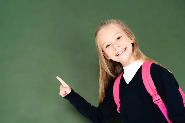 Happy schoolgirl pointing with finger at green chalkboard while smiling at camera — Stock Photo