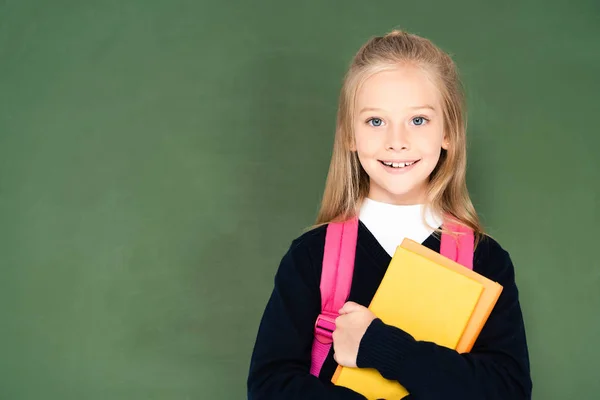 Cheerful schoolgirl holding book and smiling at camera while standing near green chalkboard — Stock Photo