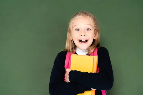 Happy schoolgirl looking at camera while holding book and standing near green chalkboard — Stock Photo