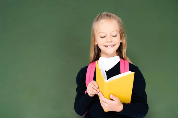 Smiling schoolgirl reading book while standing near green chalkboard — Stock Photo