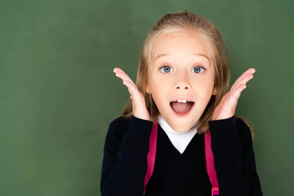 Colegiala sorprendida mirando a la cámara mientras está de pie cerca de pizarra verde - foto de stock