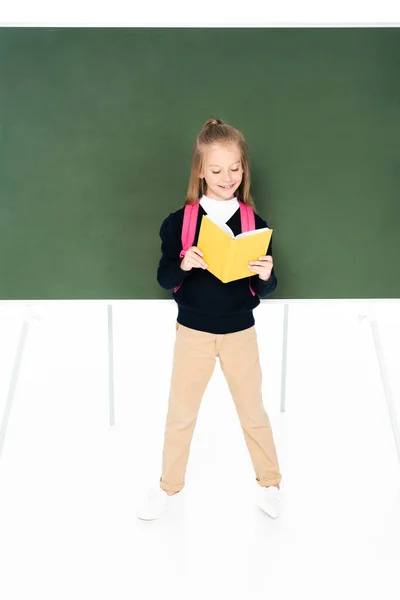 Vista completa de la colegiala leyendo libro mientras está de pie cerca de pizarra verde sobre fondo blanco - foto de stock