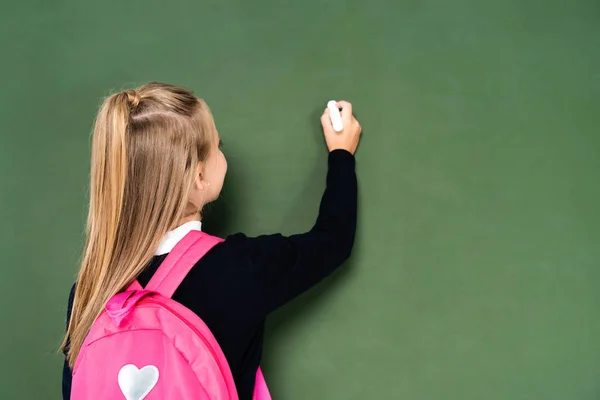 Back view of schoolgirl with pink backpack writing on green chalkboard — Stock Photo