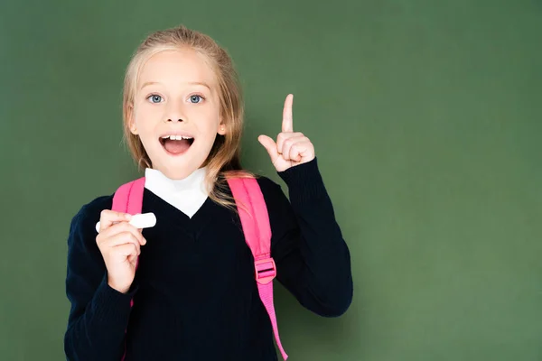 Cheerful schoolgirl holding piece of chalk and showing idea gesture while standing near green chalkboard — Stock Photo