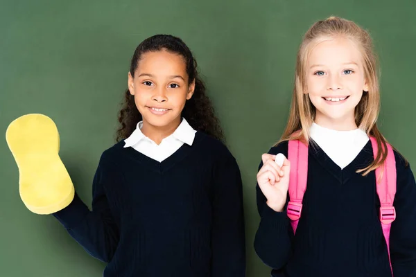 Deux écolières multiculturelles souriantes avec éponge et morceau de craie debout près du tableau vert et souriant à la caméra — Photo de stock