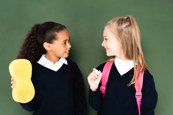 Zwei fröhliche multikulturelle Schulmädchen, die einander zuschauen, während sie neben der Tafel stehen — Stockfoto