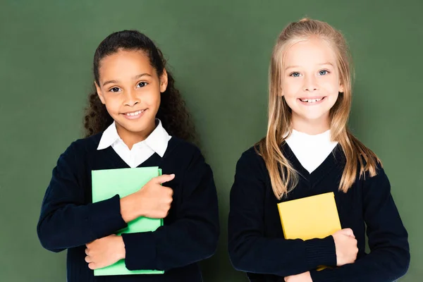 Two cute multicultural schoolgirls smiling at camera while standing near chalkboard and holding books — Stock Photo