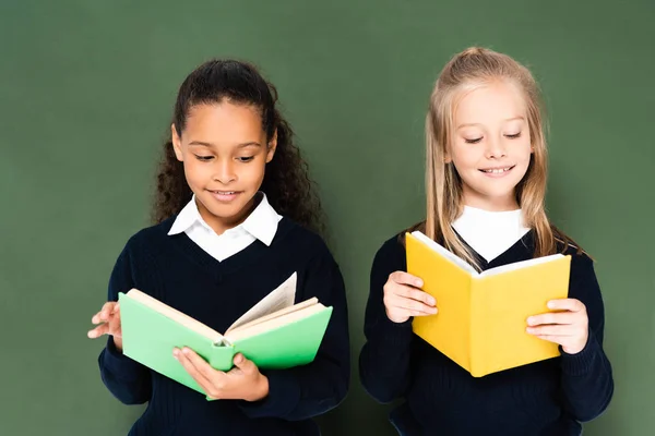 Две улыбающиеся мультикультурные школьницы читают книги, стоя рядом с зеленой доской — стоковое фото