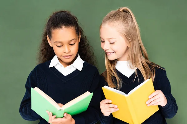 Zwei fröhliche multikulturelle Schulmädchen, die zusammen Bücher lesen, während sie neben der Tafel stehen — Stockfoto