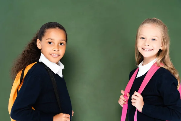 Deux écolières miltuculturelles souriantes souriant à la caméra tout en se tenant près du tableau vert — Photo de stock