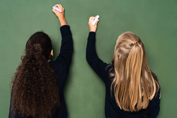 Back view of two multicultural schoolgirls writing on chalkboard with chalks — Stock Photo