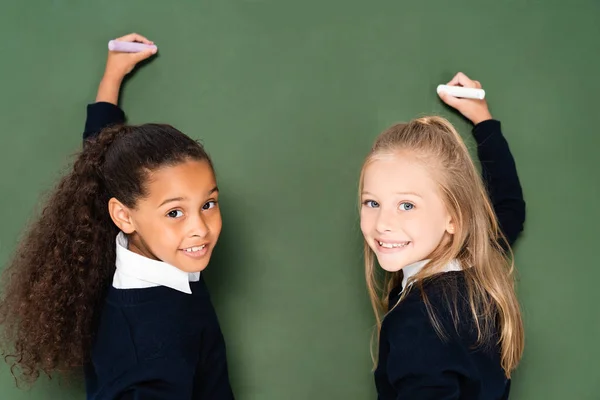 Two cheerful multicultural schoolgirls smiling at camera while writing on chalkboard — Stock Photo