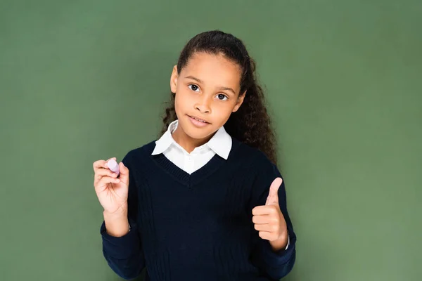 Cute african american schoolgirl showing thumb up while holding near chalkboard — Stock Photo