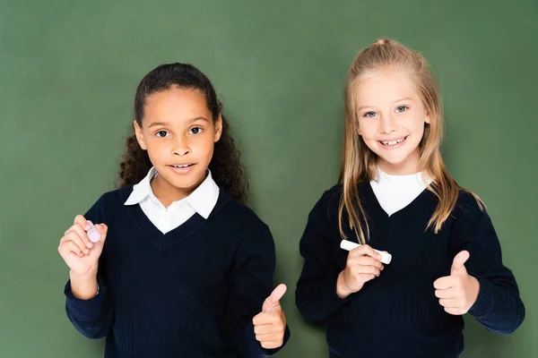 Adorable multicultural schoolgirls showing thumbs up and looking at camera while standing near chalkboard — Stock Photo