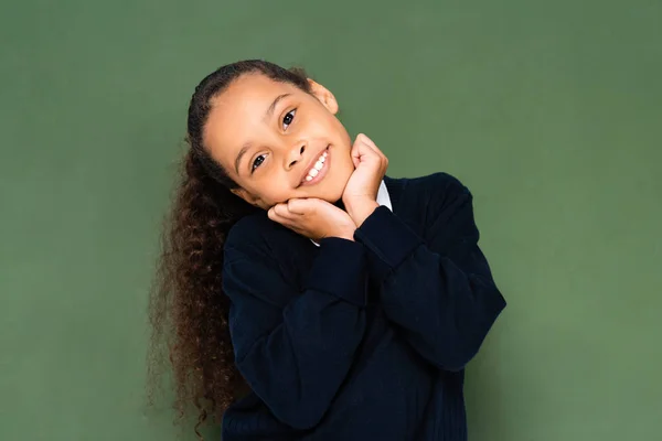 Happy african american schoolgirl holding hands near face while standing near green chalkboard — Stock Photo