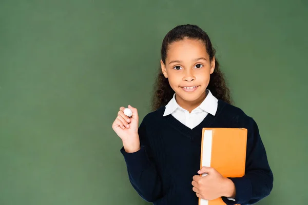 Cheerful african american schoolgirl holding piece of chalk and book while standing near chalkboard — Stock Photo