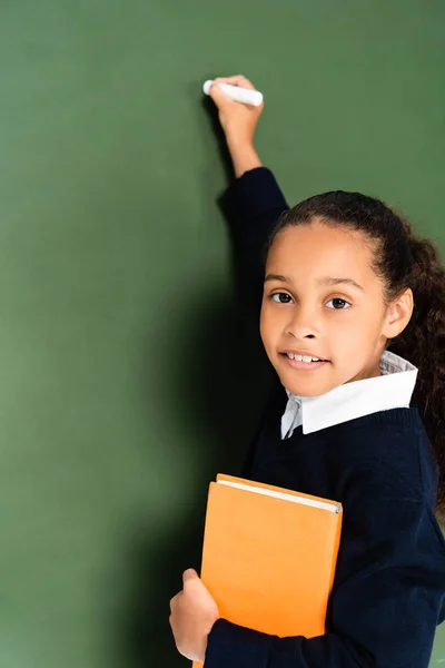 Cute african american schoolgirl writing on chalkboard while holding book and looking at camera — Stock Photo