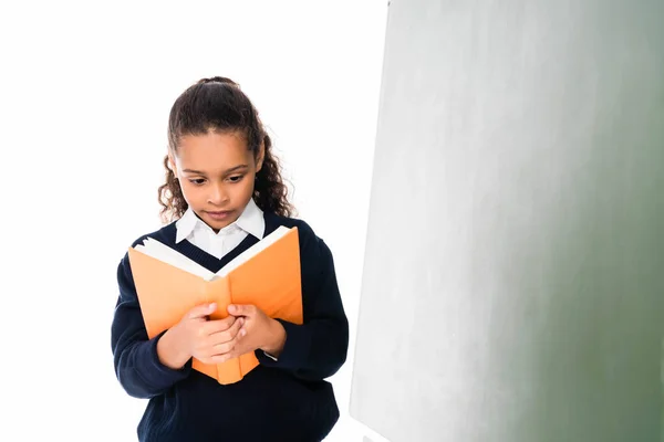 Süße afrikanisch-amerikanische Schülerin liest Buch, während sie in der Nähe von grünen Kreidetafel isoliert auf weiß steht — Stockfoto