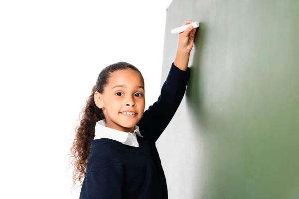 Cheerful african amercan schoolgirl writing on green chalkboard while smiling at camera isolated on white — Stock Photo