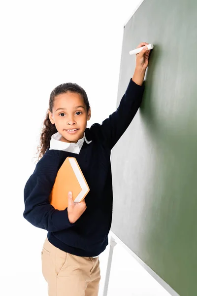 Süße afrikanisch-amerikanische Schulmädchen, die auf Kreidetafel schreibt, während sie Buch in der Hand hält und isoliert auf weiß in die Kamera schaut — Stockfoto