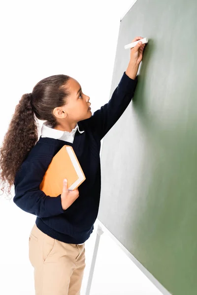 Entzückende afrikanisch-amerikanische Schülerin schreibt auf Kreidetafel, während sie Buch isoliert auf weißem Papier hält — Stockfoto