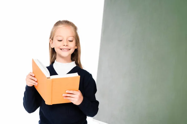 Fröhliches Schulmädchen liest Buch, während es neben der Tafel steht, isoliert auf weiß — Stockfoto