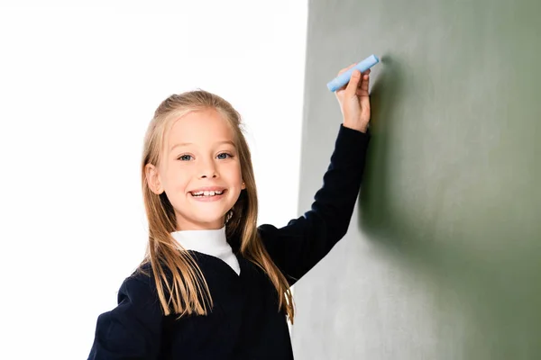 Cheerful schoolgirl smiling at camera while writing on chalkboard and smiling at camera — Stock Photo